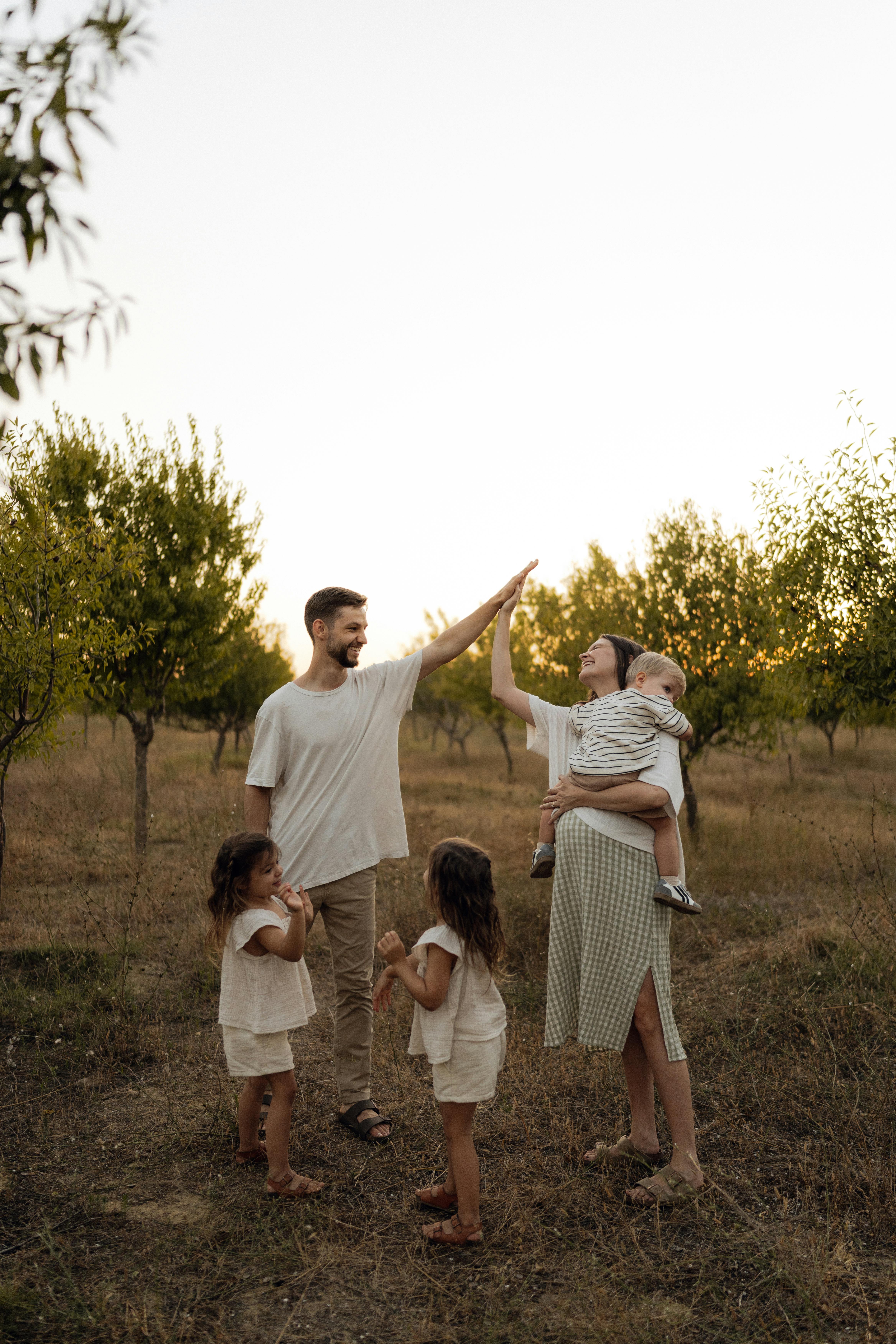 A family of five in an orchard at sunset — father and mother sharing a high-five, two young daughters and a toddler with them.